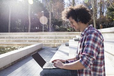 Laughing young man using laptop and headphones - ABZF01797