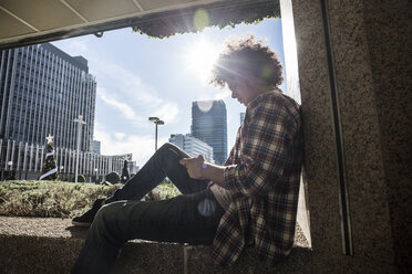 Young man sitting on wall in backlight looking at cell phone - ABZF01802