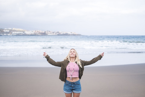 Porträt einer blonden jungen Frau, die mit geschlossenen Augen und ausgestreckten Armen am Strand steht, lizenzfreies Stockfoto