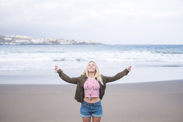 Portrait of blond young woman standing on the beach with eyes closed and arms outstretched - SIPF01349