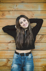 Portrait of smiling young woman standing in front of wooden wall with hands behind head - RAEF01676