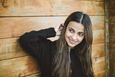 Portrait of smiling young woman in front of wooden wall watching something - RAEF01677
