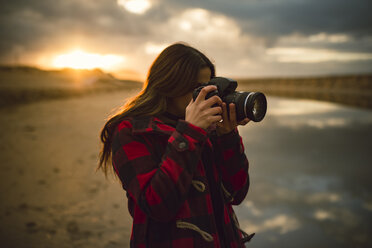 Junge Frau fotografiert am Strand mit Kamera bei Sonnenuntergang - RAEF01686