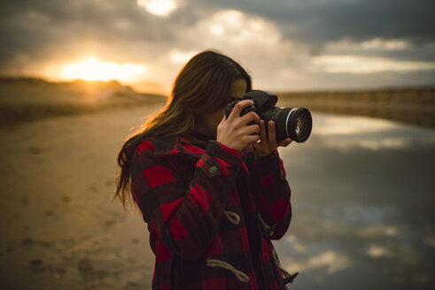 Junge Frau fotografiert am Strand mit Kamera bei Sonnenuntergang - RAEF01686
