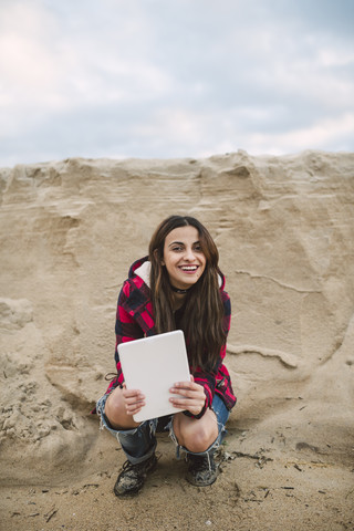 Porträt einer lächelnden jungen Frau mit Tablet, die am Strand hockt, lizenzfreies Stockfoto