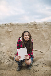 Porträt einer lächelnden jungen Frau mit Tablet, die am Strand hockt - RAEF01690