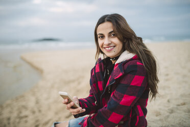 Porträt einer lächelnden jungen Frau mit Smartphone am Strand - RAEF01695