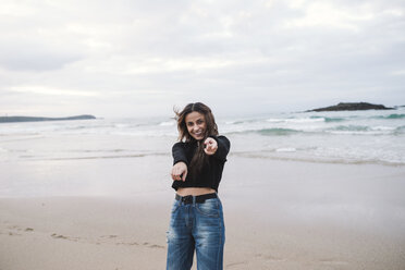 Porträt einer glücklichen jungen Frau am Strand, die auf den Betrachter zeigt - RAEF01700