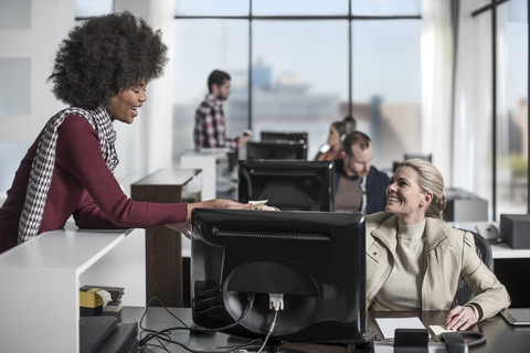 Lässige weibliche Büroangestellte, die einem Mitarbeiter hinter einem Schreibtisch mit Computer Notizen übergibt, lizenzfreies Stockfoto