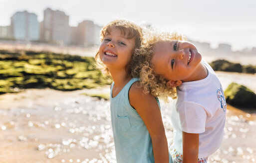 Porträt eines glücklichen kleinen Jungen und eines Mädchens, die Rücken an Rücken am Strand stehen - MGOF02841