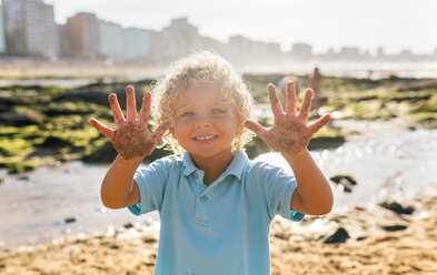 Porträt eines lächelnden kleinen Jungen, der seine sandigen Hände am Strand zeigt - MGOF02856