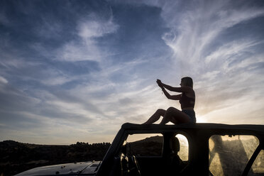 Silhouette einer Frau, die bei Sonnenuntergang auf dem Autodach sitzt und fotografiert - SIPF01386