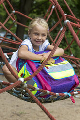 Porträt eines lächelnden kleinen Mädchens mit Schultasche, das auf einem Spielplatz in einer Nestschaukel sitzt - JFEF00830