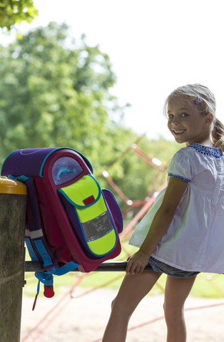 Lächelndes kleines Mädchen mit Schultasche auf dem Reck eines Spielplatzes, lizenzfreies Stockfoto