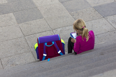 Rückenansicht eines kleinen Mädchens mit Schultasche, das auf einer Treppe sitzt und Hausaufgaben macht, lizenzfreies Stockfoto