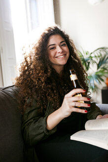 Porträt einer lächelnden jungen Frau, die mit Buch und Bierflasche auf der Couch sitzt - VABF01089