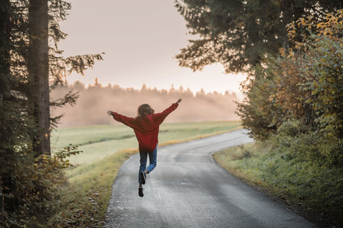 Back view of woman jumping with arms outstretched on country road in the evening - WVF00830