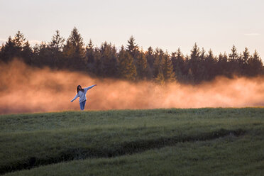 Woman with arms outstretched standing in front of haze on a meadow at sunset - WVF00831