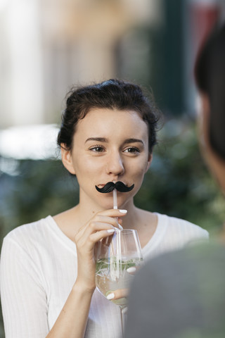 Portrait of young woman with toy moustache and cocktail at sidewalk cafe with her friend stock photo