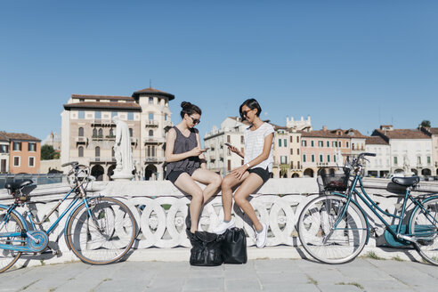 Italy, Padua, two young tourists sitting on railing looking at cell phones - ALBF00093