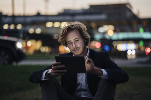 Geschäftsmann sitzt auf einer Wiese in der Abenddämmerung und benutzt ein Tablet, lizenzfreies Stockfoto