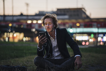 Businessman sitting on meadow at dusk with cell phone and earphones - KNSF00987