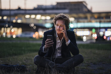 Businessman sitting on meadow at dusk with cell phone and earphones - KNSF00988