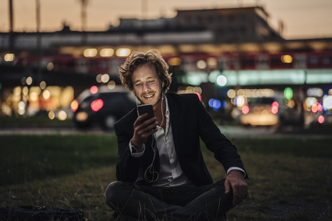 Smiling businessman sitting on meadow at dusk with cell phone and earphones stock photo