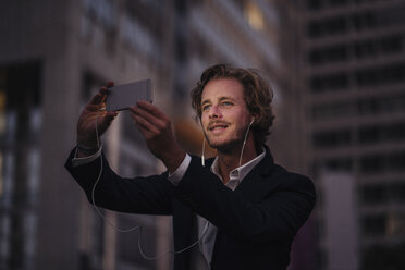 Businessman in the city at dusk with cell phone and earphones - KNSF00992