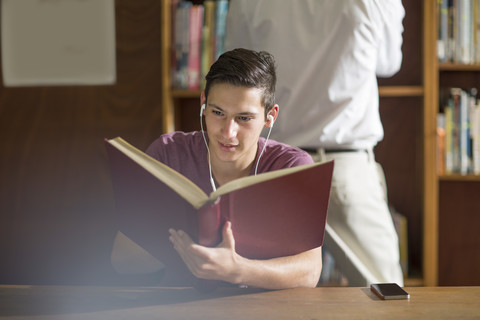 Gymnasiastin mit Kopfhörern, die in der Bibliothek ein Buch liest, lizenzfreies Stockfoto