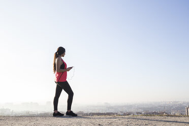 Weibliche Athletin, die mit ihrem Smartphone Musik hört, mit einer Stadtlandschaft im Hintergrund - ABZF01838