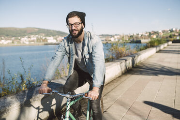 Smiling young man riding his fixie bike at the waterfront - RAEF01723