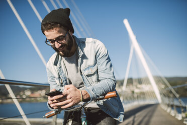 Lächelnder junger Mann mit Fixie-Fahrrad und Smartphone auf einer Brücke - RAEF01728