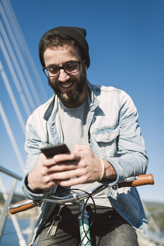 Smiling young man with fixie bike using a smartphone on a bridge stock photo