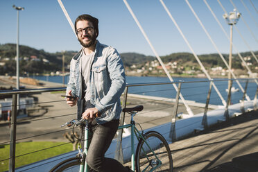 Lächelnder junger Mann mit Fixie-Fahrrad auf einer Brücke - RAEF01730