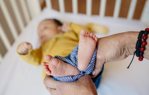 Hände der Großmutter halten die Füße des Babys in der Krippe - GEMF01474