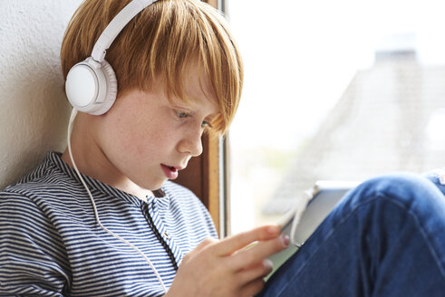 Boy sitting on window sill using digital tablet, wearing head phones - JEDF00277
