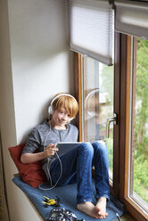 Boy sitting on window sill using digital tablet, wearing head phones - JEDF00278