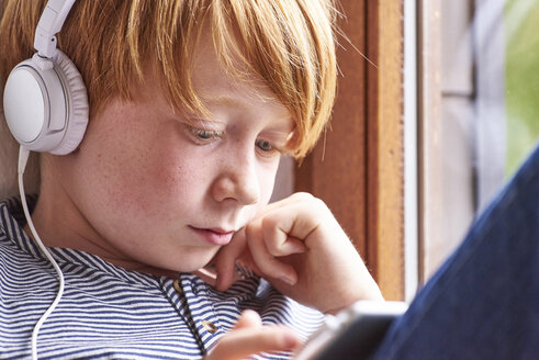 Junge sitzt auf der Fensterbank und benutzt ein digitales Tablet, er trägt Kopfhörer - JEDF00279