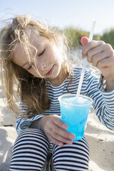 Mädchen am Strand, das ein blaues gefrorenes Getränk mit einem Strohhalm umrührt - OJF00184