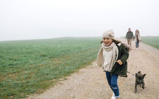 Happy girl running with her dog while her parents walking in the background on a foggy day in winter - DAPF00553