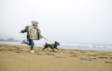 Glückliches Mädchen läuft mit Hund am Strand an einem nebligen Wintertag - DAPF00566
