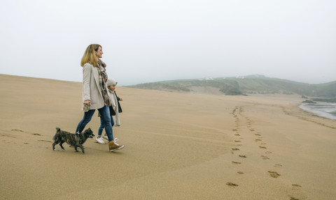 Frau, die an einem nebligen Wintertag mit Tochter und Hund am Strand spazieren geht, lizenzfreies Stockfoto