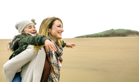 Happy woman carrying daughter piggyback on the beach in winter stock photo