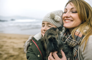 Mutter und Tochter umarmen ihren Hund am Strand im Winter - DAPF00592