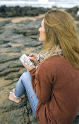 Frau mit Notebook sitzt auf Felsen am Strand im Winter - DAPF00602