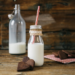 Glasflasche mit Milch und herzförmiges Schokoladengebäck auf Holz - GIOF01782