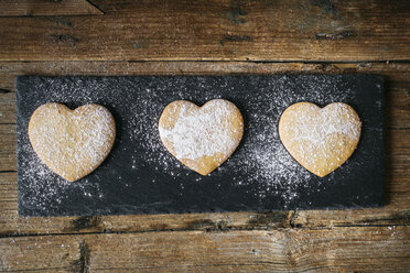 Row of three heart-shaped shortbreads sprinkled with icing sugar on slate and wood - GIOF01795