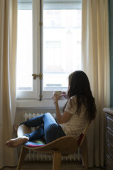 Frau mit einer Tasse Kaffee auf einem Stuhl sitzend und durch ein Fenster schauend - KKAF00361