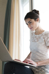 Portrait of woman sitting on window sill using laptop - KKAF00381